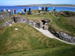 Skara Brae is situated next to Skaille Bay. When the village was built the sea would have been along the line of the mouth of the Bay. A great storm in 1859 blew away the layer of sand that had been covering the top of the village. The on-lookers are walking along the top of a sea wall that has been built to protect the site from coastal erosion.